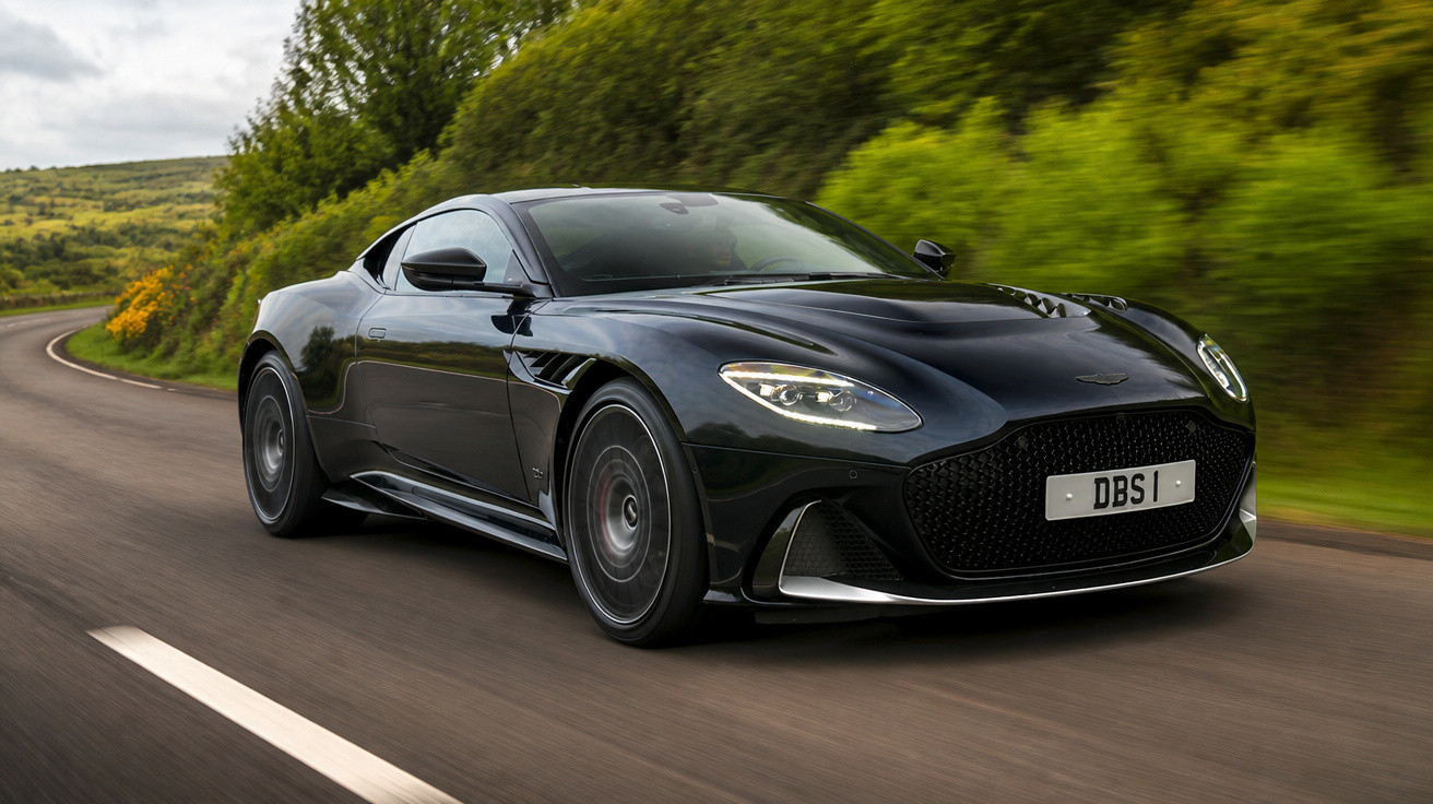 Carro esportivo preto Aston Martin DBS em alta velocidade em estrada curvas rodeada de vegetação verde.