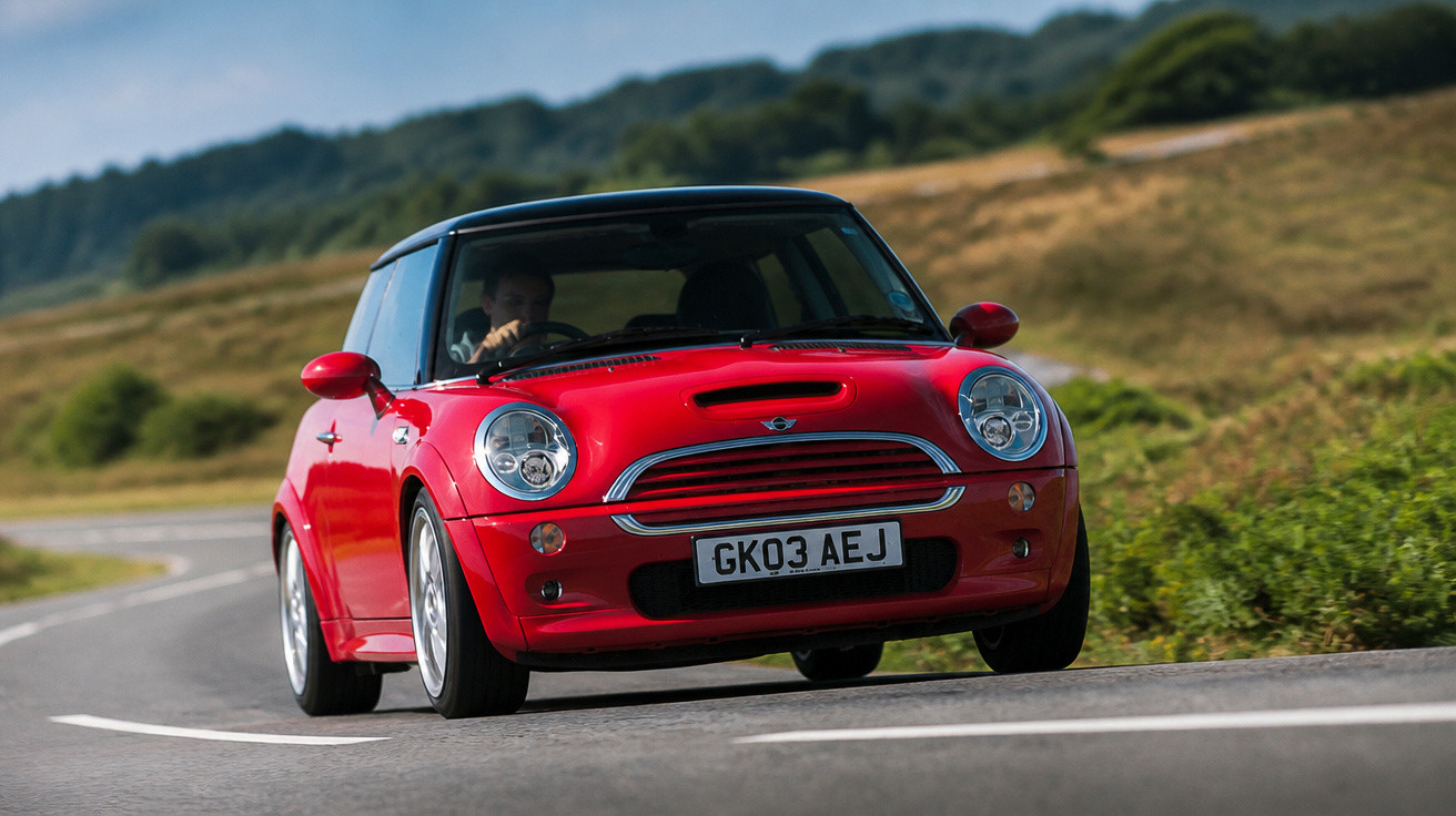 Carro Mini Cooper vermelho dirigindo em estrada sinuosa com paisagem verde ao fundo.