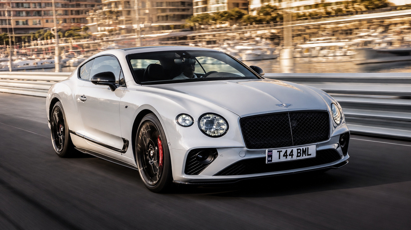Carro esportivo branco Bentley Continental GT em alta velocidade em estrada urbana com prédios ao fundo.