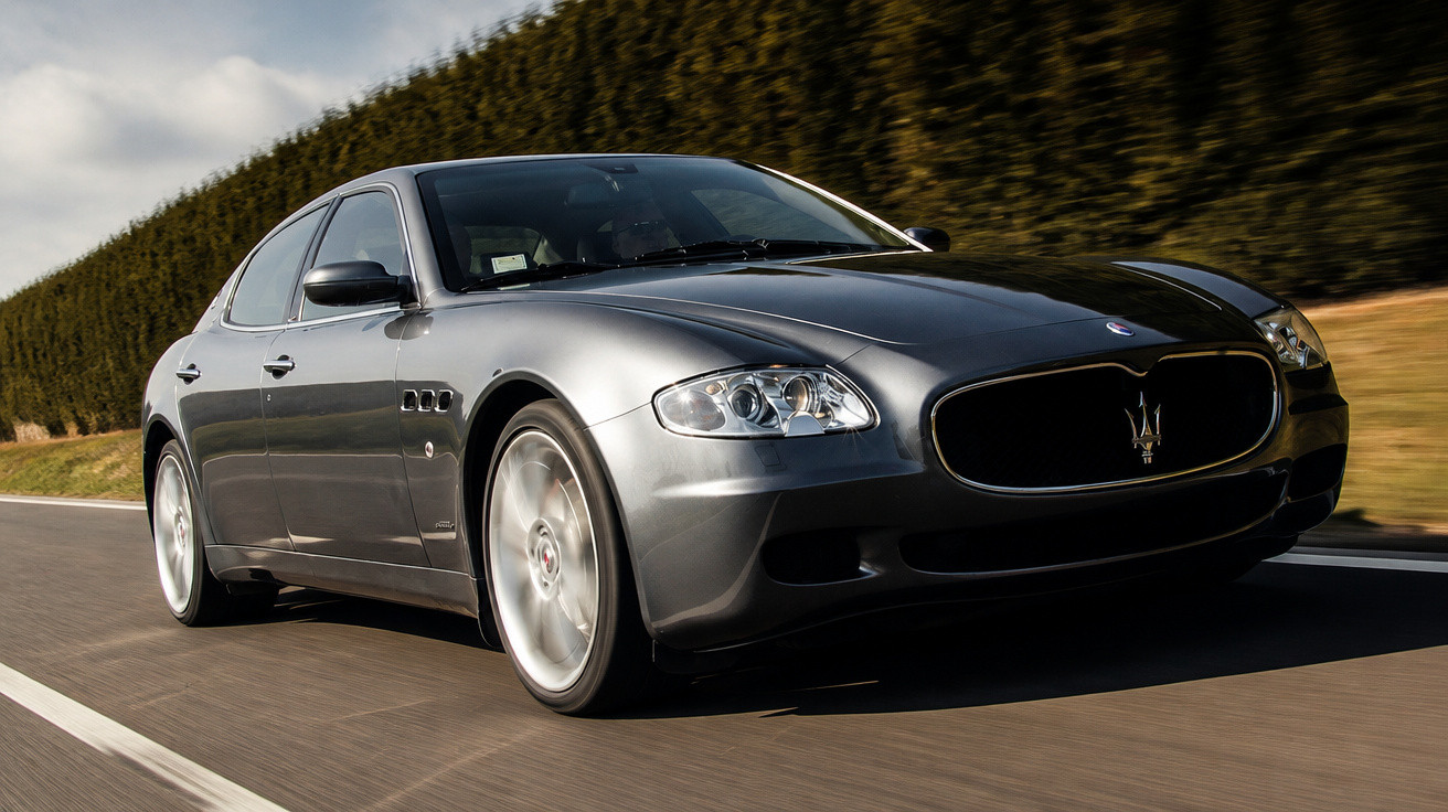 Carro esportivo cinza Maserati em movimento em estrada com vegetação ao fundo sob céu parcialmente nublado.