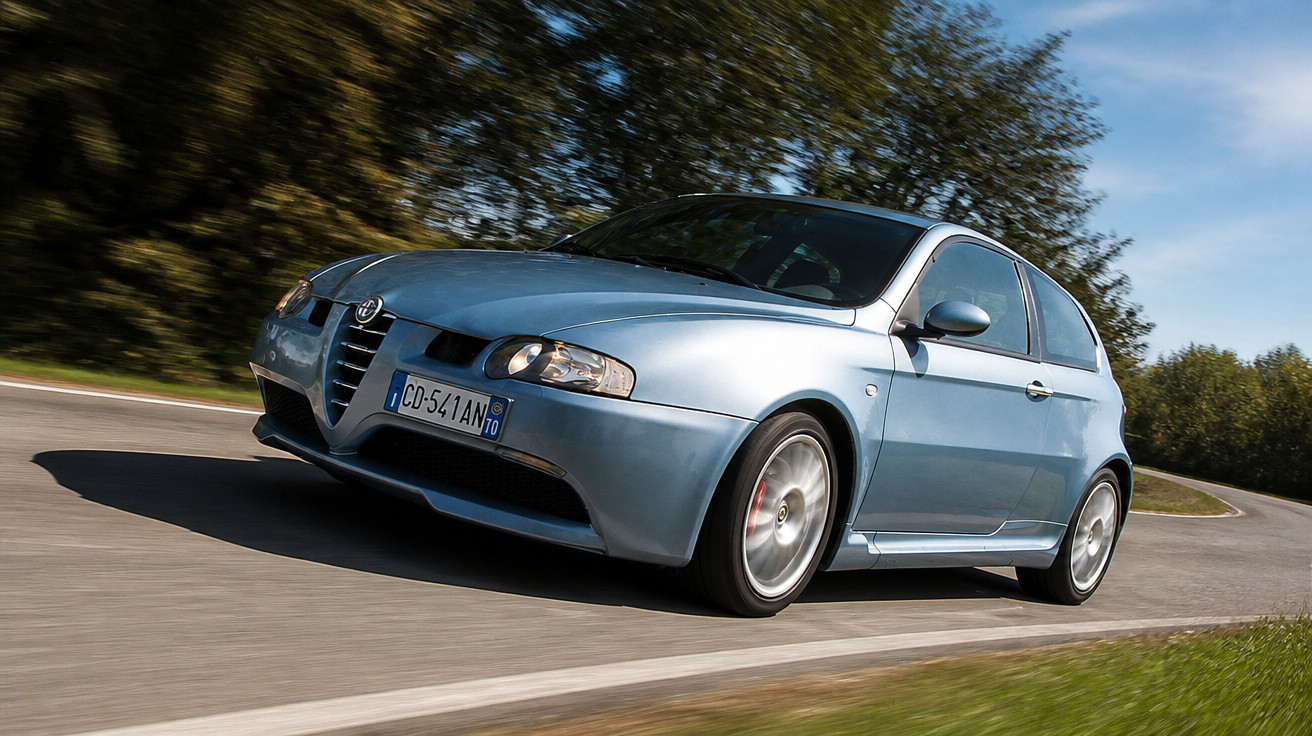 Carro Alfa Romeo cinza metálico em curva na estrada cercada por árvores e céu azul.