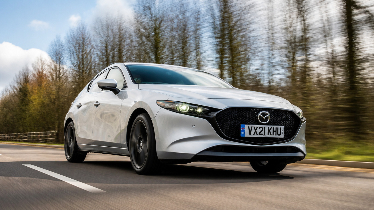 Carro prata Mazda em movimento em estrada cercada por árvores sem folhas, com céu azul ao fundo.