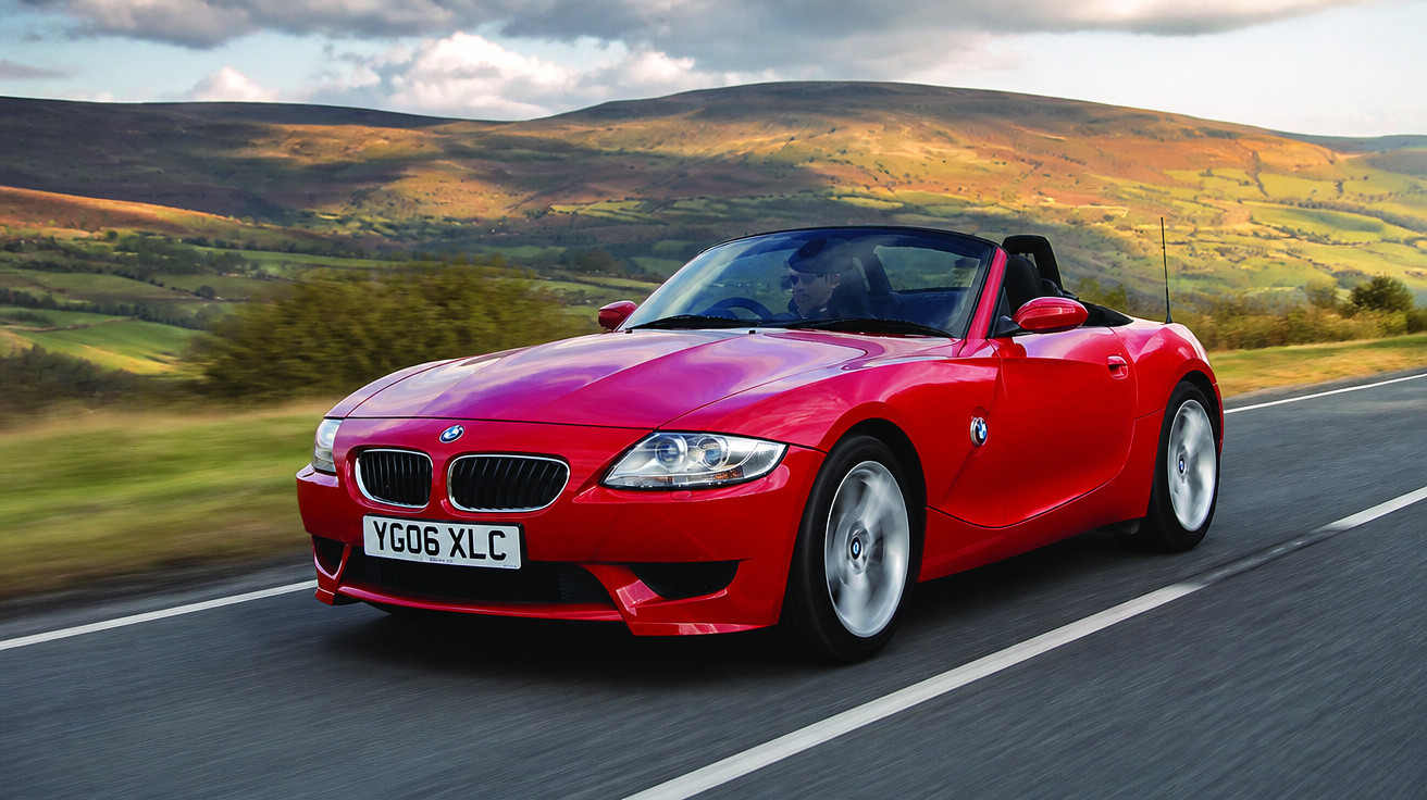 Carro conversível esportivo vermelho BMW em estrada com colinas ao fundo em dia nublado.