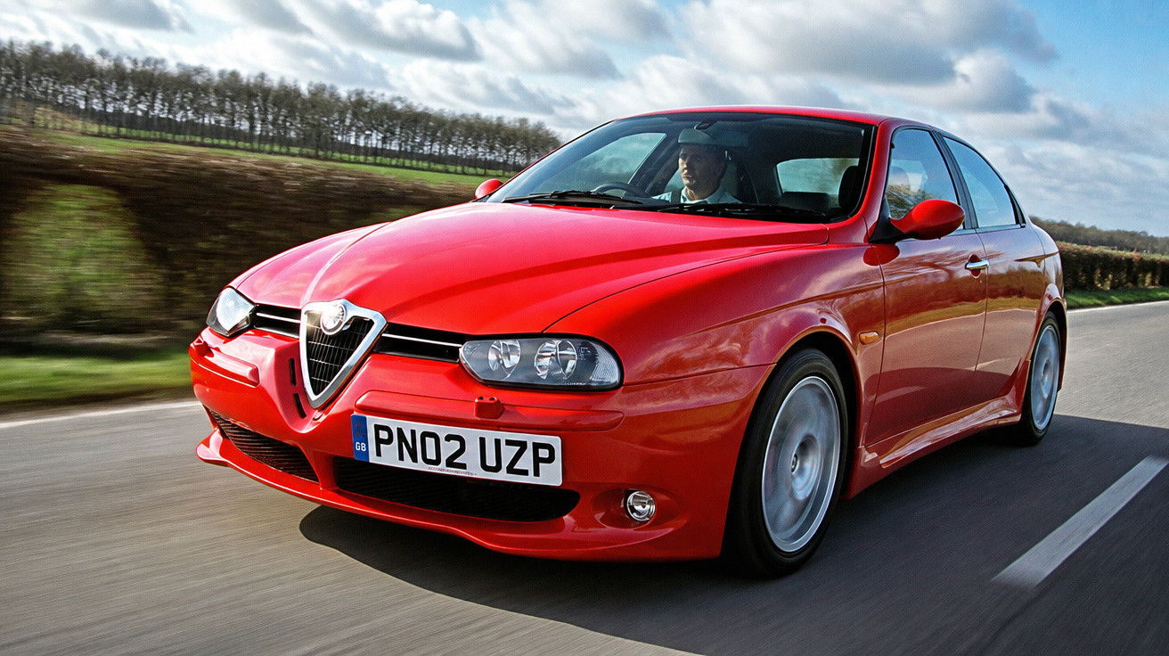 Carro esportivo Alfa Romeo vermelho em alta velocidade em estrada cercada por área verde e céu nublado.