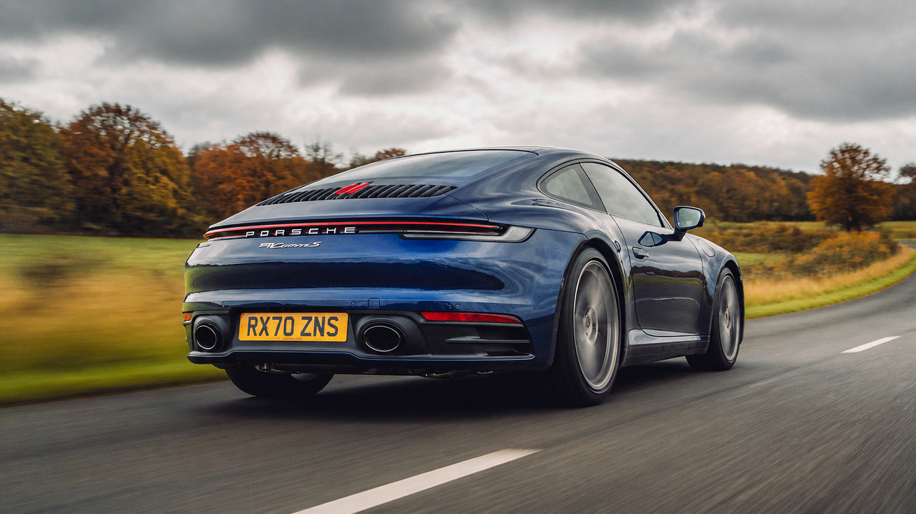 Carro esportivo Porsche 911 Carrera S azul em estrada rural com árvores ao fundo em dia nublado.