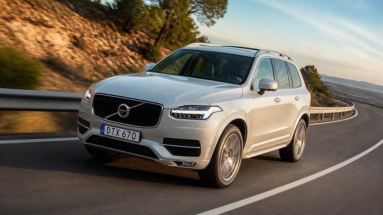SUV Volvo branco em estrada curva, com paisagem de árvores e montanhas ao fundo.