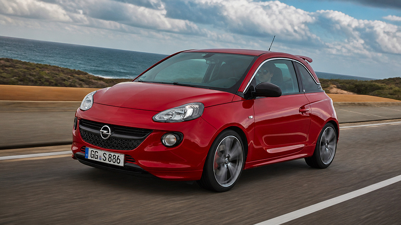 Carro esportivo vermelho Opel circulando em estrada costeira com mar ao fundo e céu nublado.