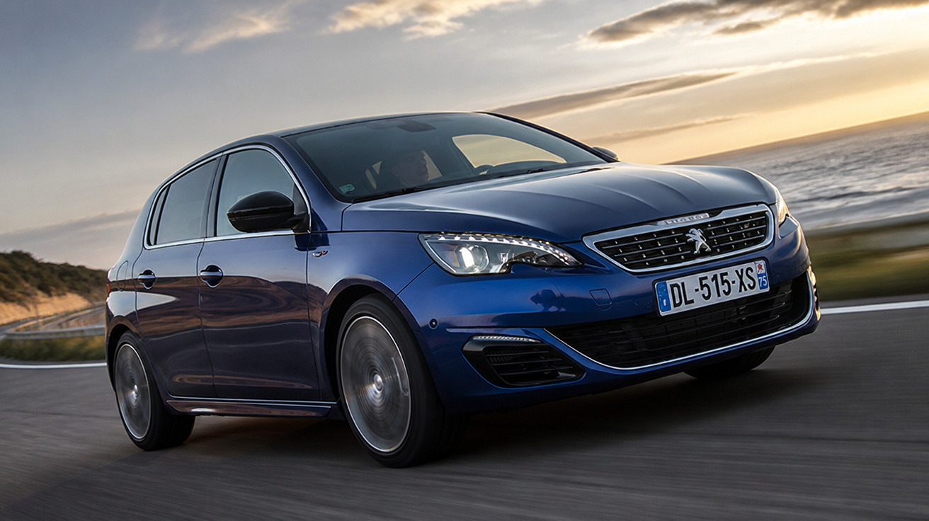 Carro azul Peugeot 308 hatch em movimento em estrada costeira ao pôr do sol.